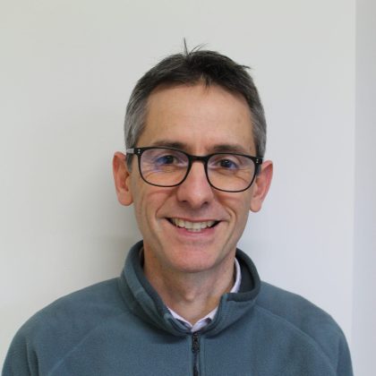 Dr Darryn Rennie smiles at the camera in front of a white background. He has short hair and is wearing glasses.