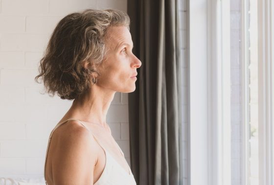 Older woman looking outside of window. She has short dark hair and is wearing a white tank top.